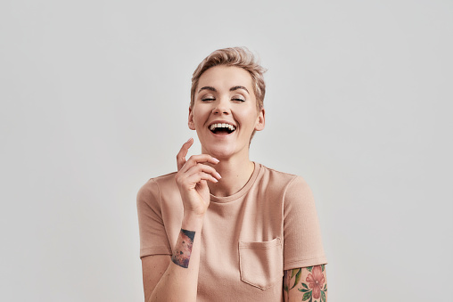 Portrait of a young tattooed woman with pierced nose and short hair in beige t shirt laughing at camera, posing with hand near face isolated over light background. Front view. Horizontal shot