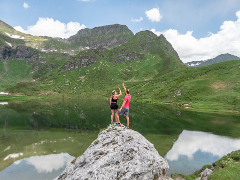 Two hikers on top of mountain enjoying lake view. Couple enjoys hiking in Summer, reaching goal, high five celebrating