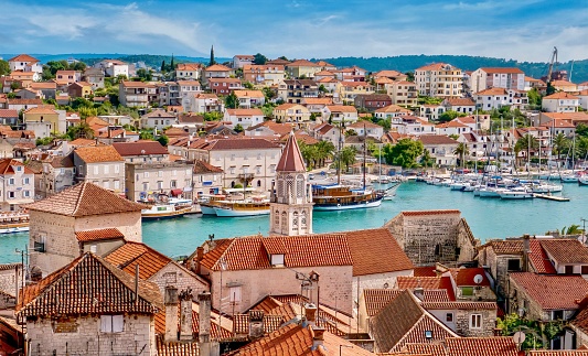 A high angle view of the Old Town of Trogir, with stone buildings and church in the foreground, while its newer buildings on the mainland are in the background divided by a channel of water containing yachts and tour boats, with a picturesque waterfront recreation area.
