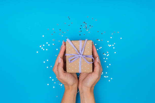 istock Women hands holding a gift or gift box decorated with confetti on a blue table top view. Flat composition for birthday or wedding. 1257901036