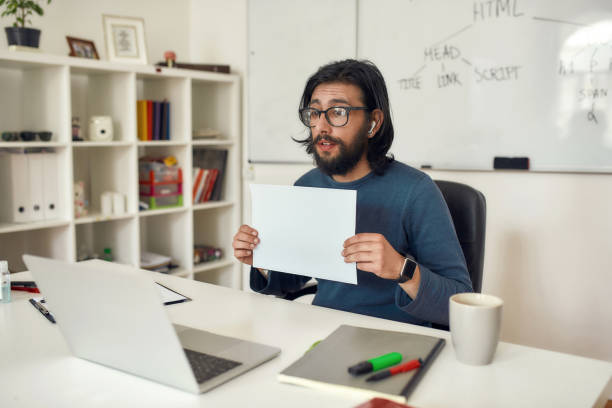 Young male computer programming tutor wearing eyeglasses showing a blank sheet of paper at web camera, using laptop and giving lesson online