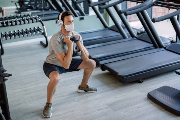 athlète masculin avec le masque de visage utilisant le poids de main tout en faisant des squats dans le club de santé. - hand weight audio photos et images de collection