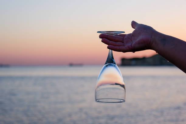 Woman's hand holding a wine glass upside down at sunset. A beautiful sea view is visible in the background. Close-up with copy space.
