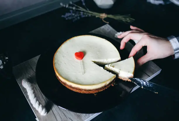 Woman cuts with knife homemade vanilla New York cheesecake on dark-violet plate on wooden tray with rope handles. Small red heart is lying on dessert. Perfect hygge Valentine's day present. Lavender