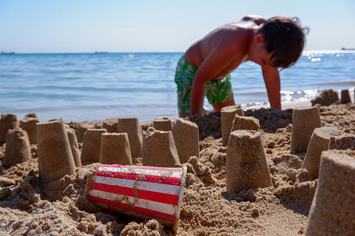 Boy making sand castles with a red and white paper cup at the beach in summer. Selective focus. Blurred background.