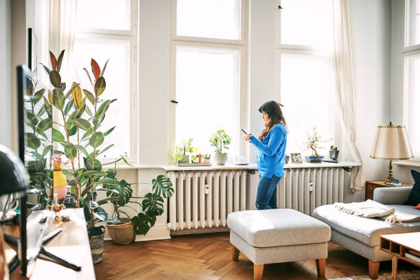 Woman using mobile phone in living room Side view of woman using smart phone. Female is standing by window in living room. She is at home. radiator heater stock pictures, royalty-free photos & images