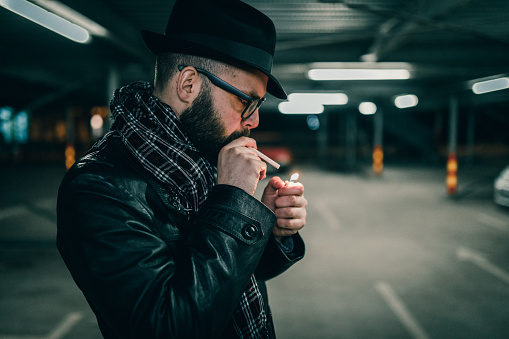 Young man standing in the parking lot in the night and lighting a cigarette