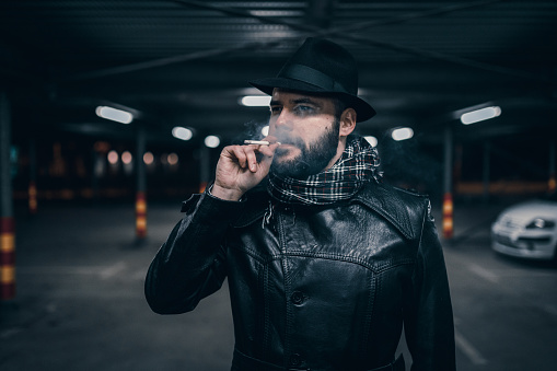 Man wearing a hat smoking a cigarette in the parking lot in the night