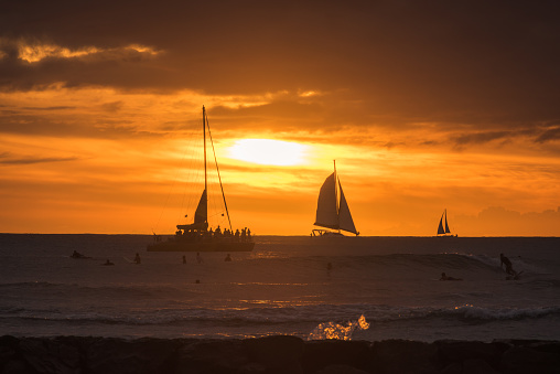 Honolulu, USA - November 10, 2017: A vivid sunset sail boats and surfers on the horizon just off Waikiki beach.