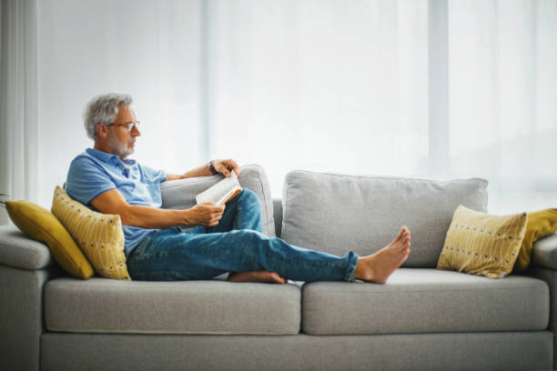 uomo maturo che legge un libro sul divano. - comfortable relaxation sofa men foto e immagini stock