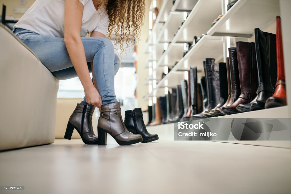 Young woman choosing and buying shoes in boutique. Beautiful young woman with brown curly hair choosing and and trying out one pair of shoes in shoe store. She is casually dressed. Shoe Store Stock Photo