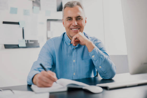 istock handsome gray-haired man sits in his office writing notes to his agenda while working on business project looks satisfied, work routine concept 1257921318