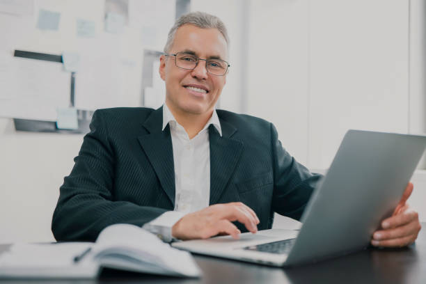 istock handsome gray-haired businessman in elegant suit sits in office looking excited while working in lap top on new business project , money making motivation concept 1257921351