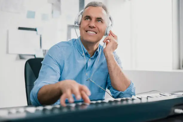 Photo of handsome gray-haired smiling man sits in his music studio playing keyboard piano wearing earphones looking excited and happy, music record concep, art