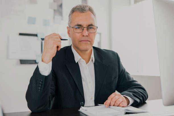 istock handsome gray-haired businessman in elegant suit sits in his office while working on business project looks confident, takes notes to his agenda, work routine concept 1257921382