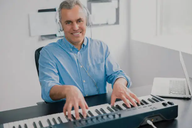 Photo of handsome gray-haired smiling man with earphones sits in music studio playing keyboard piano enjoying music looking happy, music record concep, art of composition