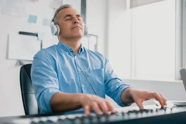 Photo of handsome gray-haired smiling man with headphones sits in music studio playing keyboard piano enjoying music looking excited and happy, music record concep, art of composition