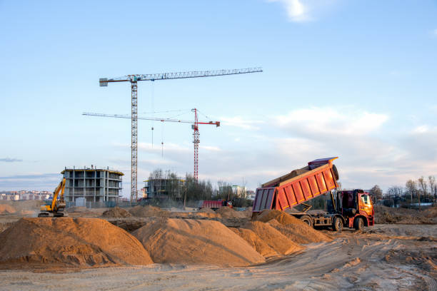 istock Dump truck unloading earth sand for road construction or for foundation work. Work of tower crane at construction site on blue sky background. Tall house renovation project, government programs 1257922750