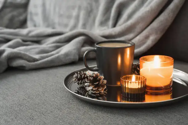 Photo of Aroma candles of orange color, coffee in a black mug and decorative pine cones served on a metal tray. Autumn or winter atmosphere