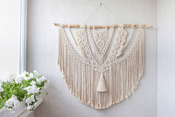istock Handmade macrame. 100% cotton wall decoration with wooden stick hanging on a white wall.   Macrame braiding and cotton threads.  Female hobby.  ECO friendly modern knitting DIY natural decoration concept in the interior 1257929658