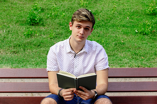Young male reading a book while sitting on a bench in a park