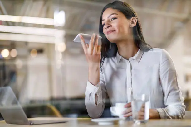 Beautiful businesswoman talking to virtual assistant on her phone while working in the office. The view is through glass.