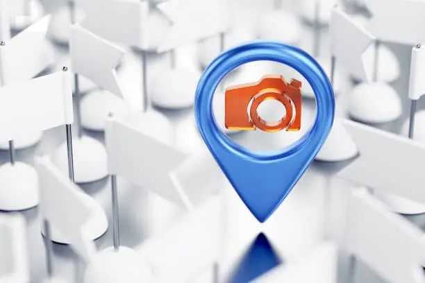 Blue colored location pointer with a camera icon in center of it in surrounding of identical white pennants which are randomly arranged on reflective white surface. 3D rendering graphics on the theme of GPS Technology.