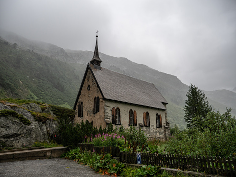 Low angle view of beautiful traditional Swiss church in Spring/ Summer. Chapel with purple flowers in front, low clouds mystic ambiance