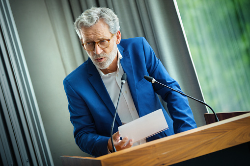 Closeup front view of an early 60's company executive having a public addressing in a board room. He's talking about recovery strategy after coronavirus economy collapse.