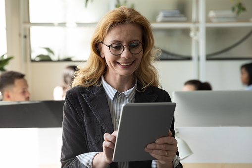 Happy attractive mature businesswoman using tablet and getting good news. Smiling middle age woman with glasses using mobile modern technology for online business chat.