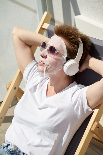 Happy pretty young girl with tissue facial mask on face sitting on the balcony and listening to music on headphones.