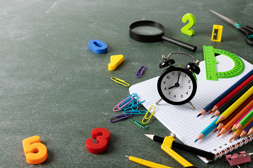 Back to school and education concept with small alarm clock, paper notebook and school supplies on blackboard background.