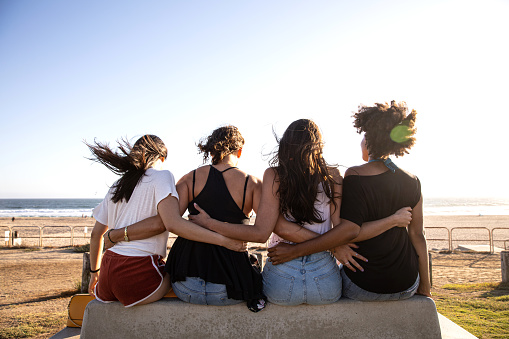Four friends hugging sitting on bench in front of a beach in Californi while traveling together.