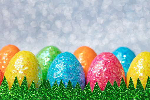 Close-up of multi colored Easter eggs on a gray sparkling defocused background. Artificial grass made of sparkling green paper. Shallow depth of field, space for copy.