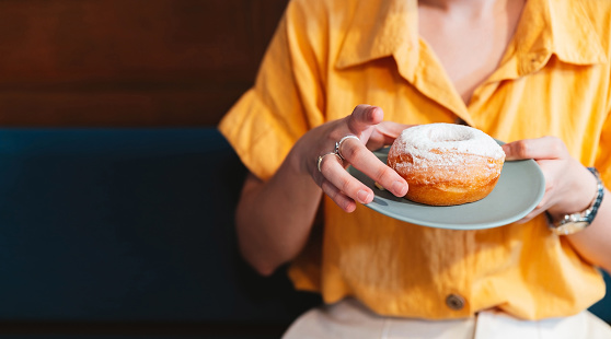 Woman wearing yellow shirt holding and showing a pale green plate of sugar glazed donut in a modern cafe. Enjoyment female lifestyle. Focus on hand and donut.