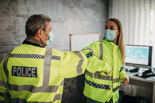 Senior male and young female police officers with protective face masks greeting with elbows in the office, avoid handshake.