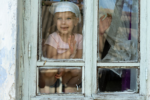 little girl looks out the window of an old village house, view through the glass