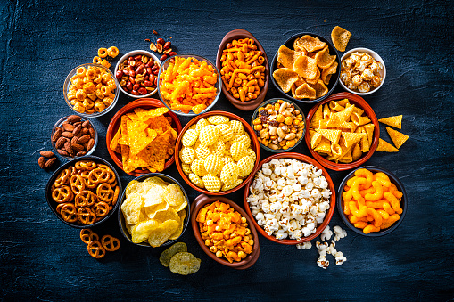 Party food: assortment of salty snacks in bowls shot from above on on dark slate table. The composition includes potato chips, popcorn, corn bugles, pretzels, peanut, cheese sticks, almonds, nachos and others. Predominant colors are yellow and black. High resolution 42Mp studio digital capture taken with SONY A7rII and Zeiss Batis 40mm F2.0 CF lens