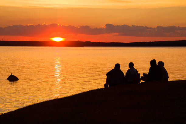 Petrozavodsk, Russia - July 18TH, 2020: Four unknown human silhouettes are sitting on the shore and watching a beautiful sunset