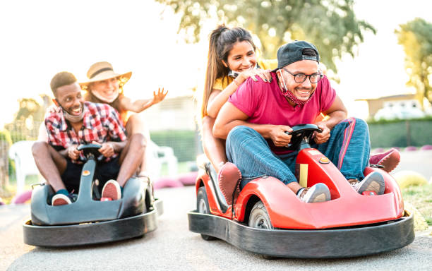 Milenial friends having fun at children playground on go kart race - Young people with face mask competing on mini car racing - New normal lifestyle concept with focus on right guy - Backlight filter Milenial friends having fun at children playground on go kart race - Young people with face mask competing on mini car racing - New normal lifestyle concept with focus on right guy - Backlight filter go carting stock pictures, royalty-free photos & images