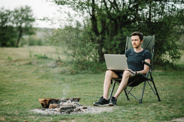 istock Man working on laptop on nature. Young freelancer relaxing in forest. Remote work, outdoor activity in summer. Travel, hiking, technology, tourism, people concept - man sitting on chair outdoors. 1258004982