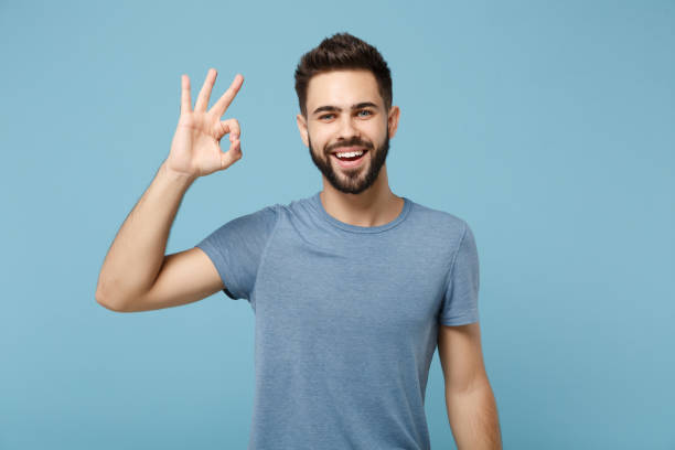 istock Young funny smiling handsome man in casual clothes posing isolated on blue wall background, studio portrait. People sincere emotions lifestyle concept. Mock up copy space. Showing OK gesture. 1258006816