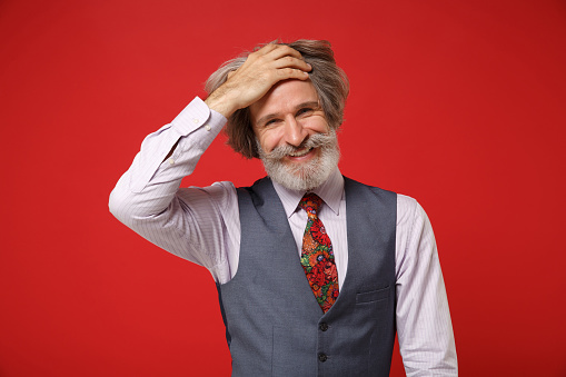 Smiling elderly gray-haired mustache bearded man in classic shirt vest and colorful tie posing isolated on red background in studio. People lifestyle concept. Mock up copy space. Putting hand on head