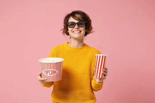 Smiling young woman girl in 3d imax glasses posing isolated on pink background. People in cinema, lifestyle concept. Mock up copy space. Watching movie film, holding bucket of popcorn, cup of soda