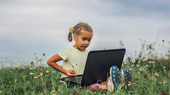 Cute little kid, pupil, sitting on the green grass in mountains and using a laptop for studying and communication online, summer outdoor, social network, gadget and technology concept