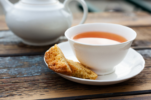 Part view of a cup of golden tea, with a home-baked almond biscuit on the saucer. White porcelain cup and saucer on a rustic wooden table surface with a white teapot in the background. Outdoor shot with natural light.