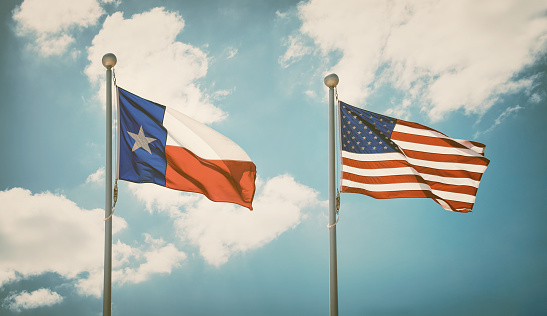The state flag of Texas and American flag waving in the wind on flagpoles. Blue sky background and white clouds. Vintage filter effects.
