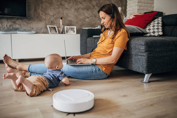 istock Young mother working on laptop at home while robotic vacuum cleaner cleaning floor 1258021593