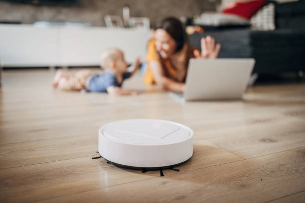 Mother working on laptop at home while robotic vacuum cleaner cleaning floor Young mother working on laptop at home while her baby boy playing next to her and robotic vacuum cleaner cleaning floor. smart home family stock pictures, royalty-free photos & images