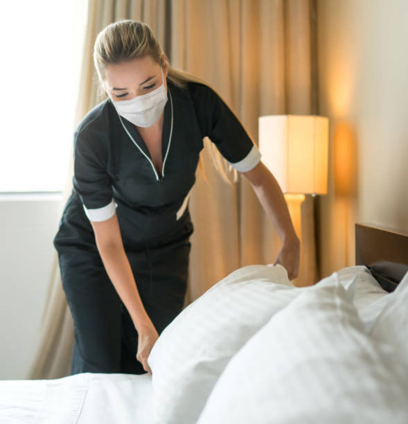 Housekeeper doing the bed wearing a facemask while working at a hotel Housekeeper doing the bed wearing a facemask while working at a hotel - Pandemic lifestyle concepts housekeeping staff photos stock pictures, royalty-free photos & images